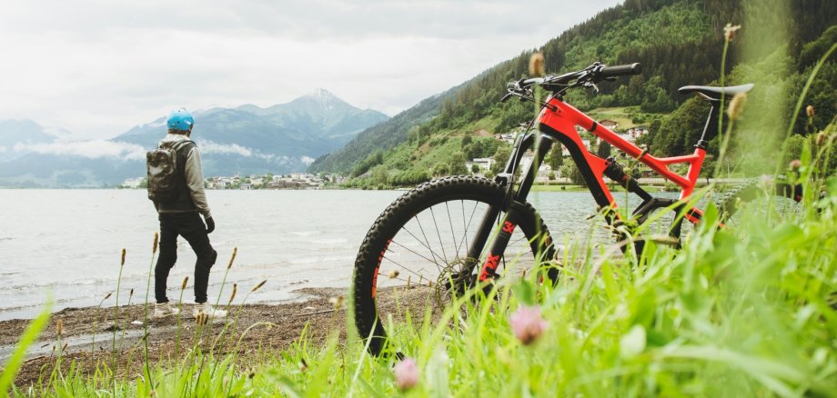Mountainbike Natur Fahrradfahrer vor einem Panorama mit See und Bergen. Sein Mountainbike steht im Vordergrund des Bildes.