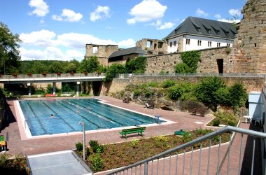 Blick auf ein Schwimmerbecken an einem sommerlichen hellen Tag. Oberhalb des Beckens auf der rechten Seite des Bildes ist eine Burgmauer zu erkennen. Im hinteren teil ein teil der Burg weiß verputzt. 
