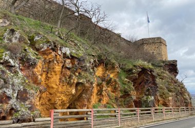 Felswand mit orangefarbenen Verwitterungen und Bewuchs, darüber ein runder Turm mit Fahne, davor eine Straße mit Holzgeländer.