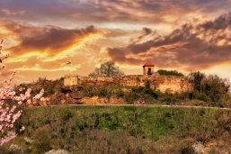 Blick auf eine Burgmauer mit einem Türmchen. Im Vordergrund ein grün bewachsener Hang. Der Himmel ist leicht bewölkt im Abendrot. Vorne links im Vordergrund ist ein Mandelblütenzweig zu sehen.