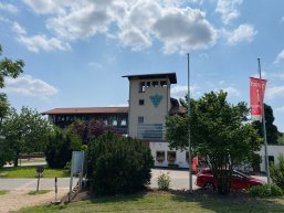Mehrstöckiges Gebäude mit rotem Ziegeldach, Weintrauben-Logo an der Fassade und roter Fahne neben einem Baum im Vordergrund.