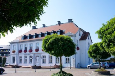 Weißes barockes Schloss mit rotem Walmdach, mehreren Dachgauben und Blumenkästen an den Fenstern, davor kleine Bäume und geparkte Autos.