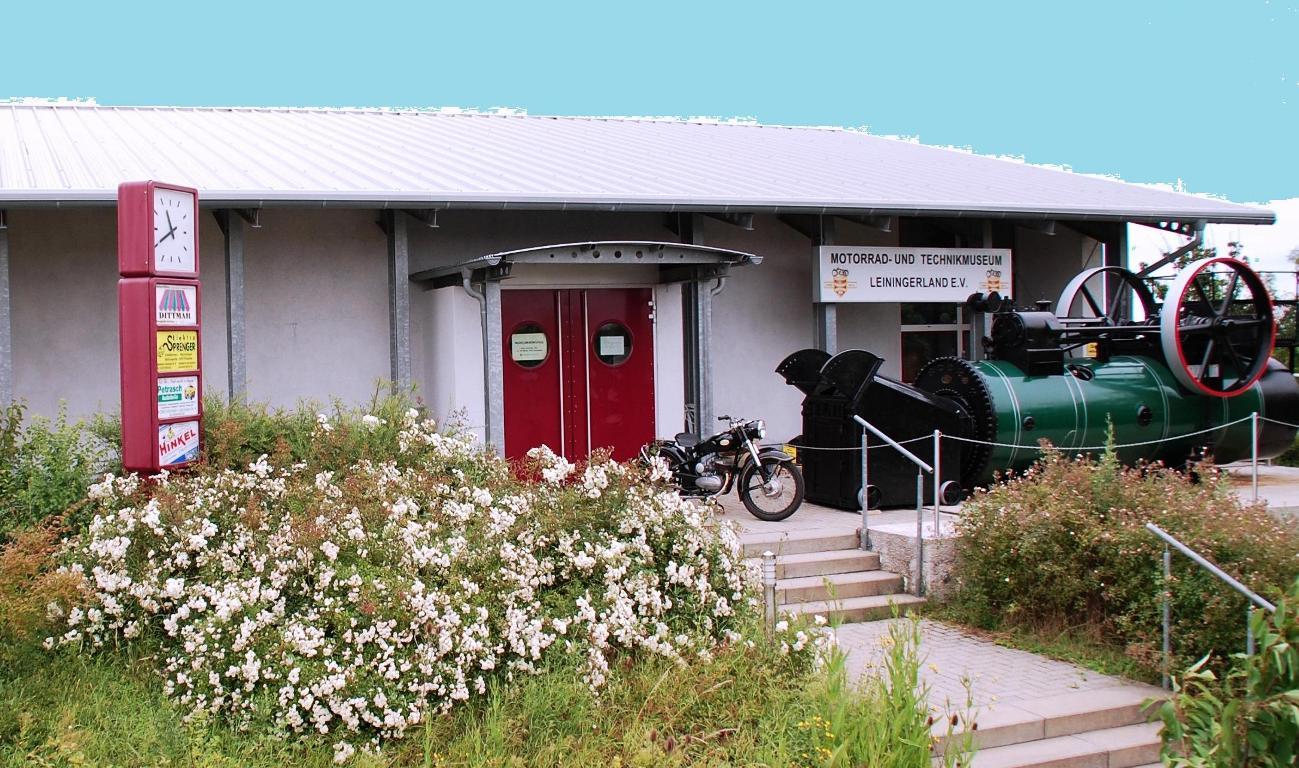 Eingang zum Motorrad- und Technikmuseum mit roten Doppeltüren, einem alten Motorrad und einer großen grünen Dampfmaschine rechts.