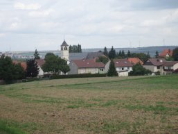 Blick auf ein Dorf mit Kirche, mehreren Häusern und Windrädern am Horizont unter bewölktem Himmel.