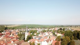 Luftaufnahme von Kirchheim mit Kirchturm, roten Dächern, Wohnhäusern und umgebenden Feldern unter klarem Himmel.