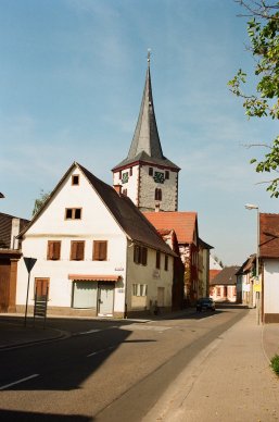Straße in Laumersheim mit Fachwerkhäusern und einem Kirchturm mit spitzem Schieferdach unter blauem Himmel.