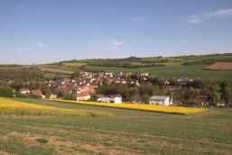 Blick auf ein Dorf mit roten Dächern, umgeben von grünen und gelben Feldern unter blauem Himmel.