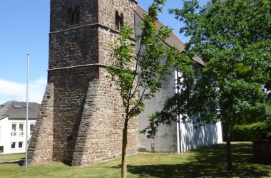 Kirchturm aus grobem Stein mit Spitzdach neben einem jungen Baum auf einer Wiese unter blauem Himmel.