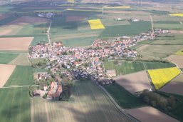 Luftaufnahme eines Dorfes mit roten Dächern, umgeben von landwirtschaftlichen Feldern in verschiedenen Grüntönen und einigen gelben Blühflächen.