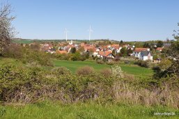 Dorf mit roten Ziegeldächern, Kirche, zwei Windrädern im Hintergrund und grünen Feldern im Vordergrund.