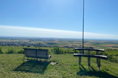 Zwei gebogene Holzbänke auf einer Wiese mit weitem Blick über Felder und Windräder am Horizont.