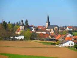 Blick auf ein Dorf mit zwei Kirchen, mehreren Häusern mit roten Dächern und Feldern im Vordergrund.