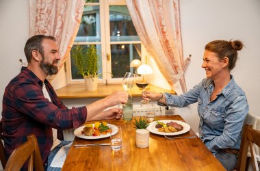Zwei Personen sitzen an einem Holztisch, stoßen mit Weingläsern an, auf dem Tisch stehen Teller mit Essen, eine Wasserflasche und eine kleine Pflanze.