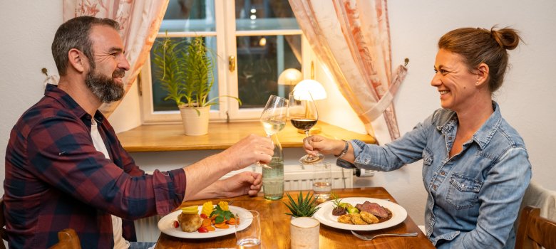 Zwei Personen sitzen an einem Holztisch, stoßen mit Weingläsern an, auf dem Tisch stehen Teller mit Essen, eine Wasserflasche und eine kleine Pflanze.
